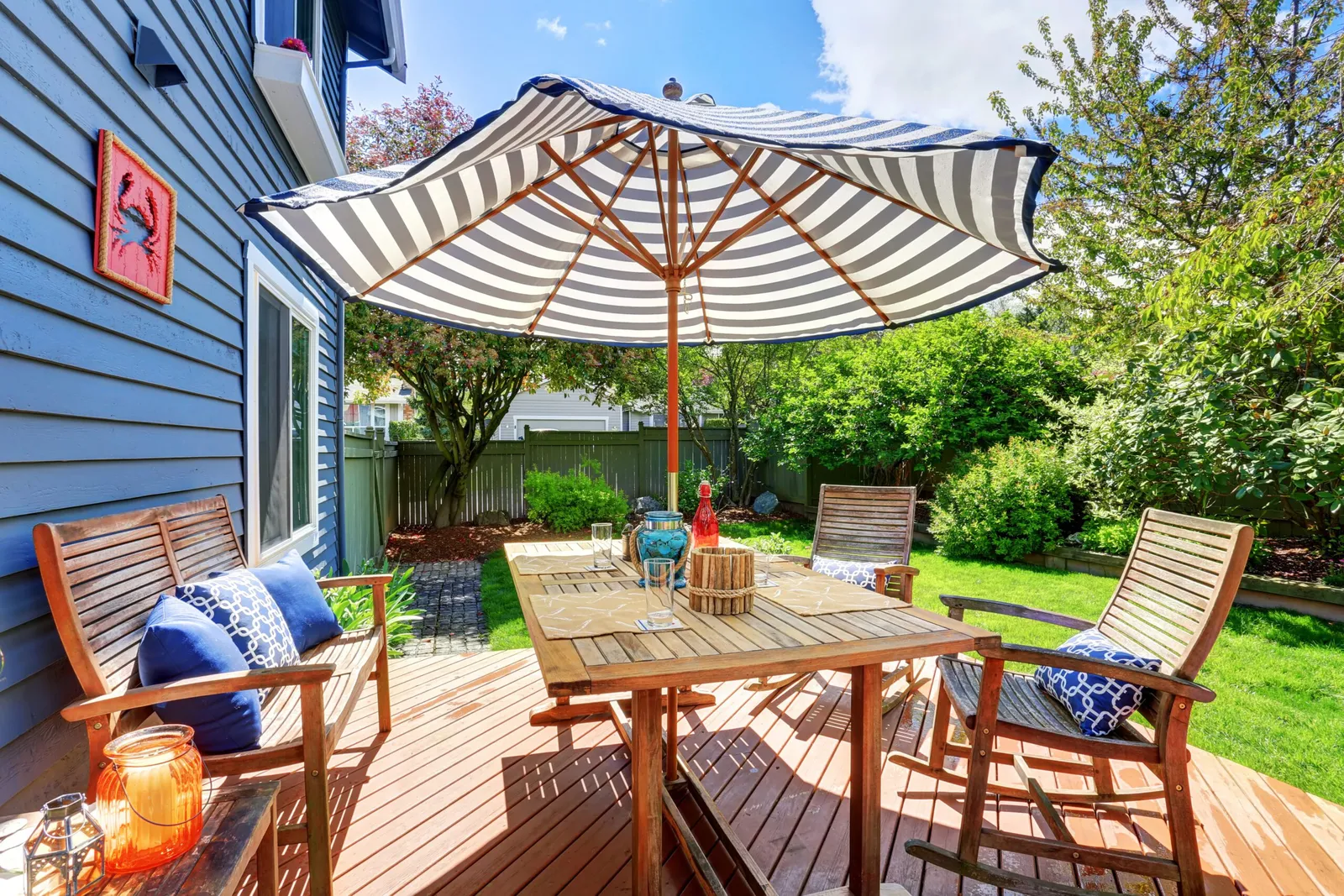 A spacious backyard deck with a striped umbrella, wooden dining table, and cozy seating area, surrounded by greenery and sunlight, creating a warm and welcoming atmosphere.