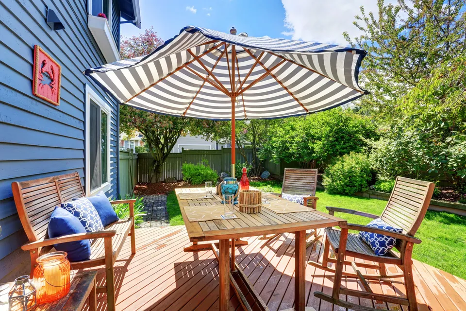 A spacious backyard deck with a striped umbrella, wooden dining table, and cozy seating area, surrounded by greenery and sunlight, creating a warm and welcoming atmosphere.