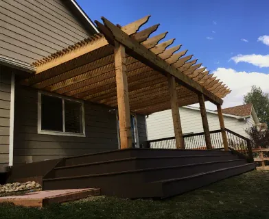 A ground-level composite deck attached to a beige house, featuring a large wooden pergola with exposed beams and angled rafters, creating a shaded outdoor area ideal for relaxing or entertaining.
