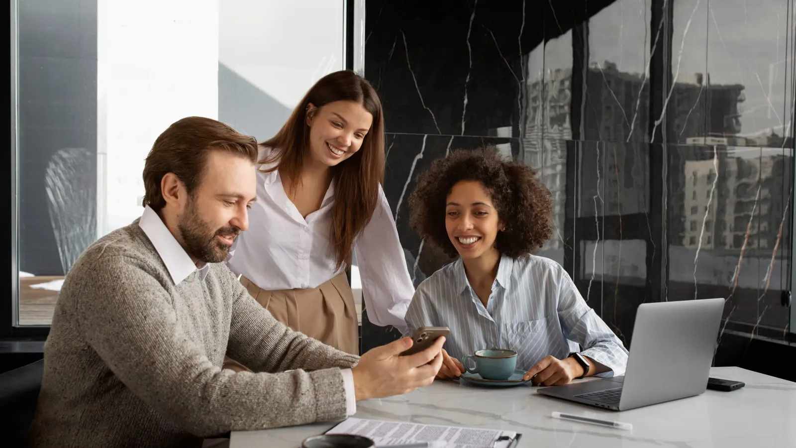 Equipe de vendas reunida à mesa olhando juntos o celular e o notebook